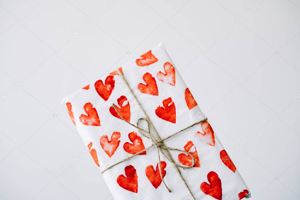 a cute gift in a woman's hand wrapped in homemade wrapping paper with red hearts tied with jute thread for Valentine's day on a white background