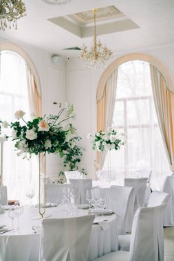 Çiçekli düğün masaları, narin beyaz çiçekler ve beyaz salonda taze yeşillik dalları.