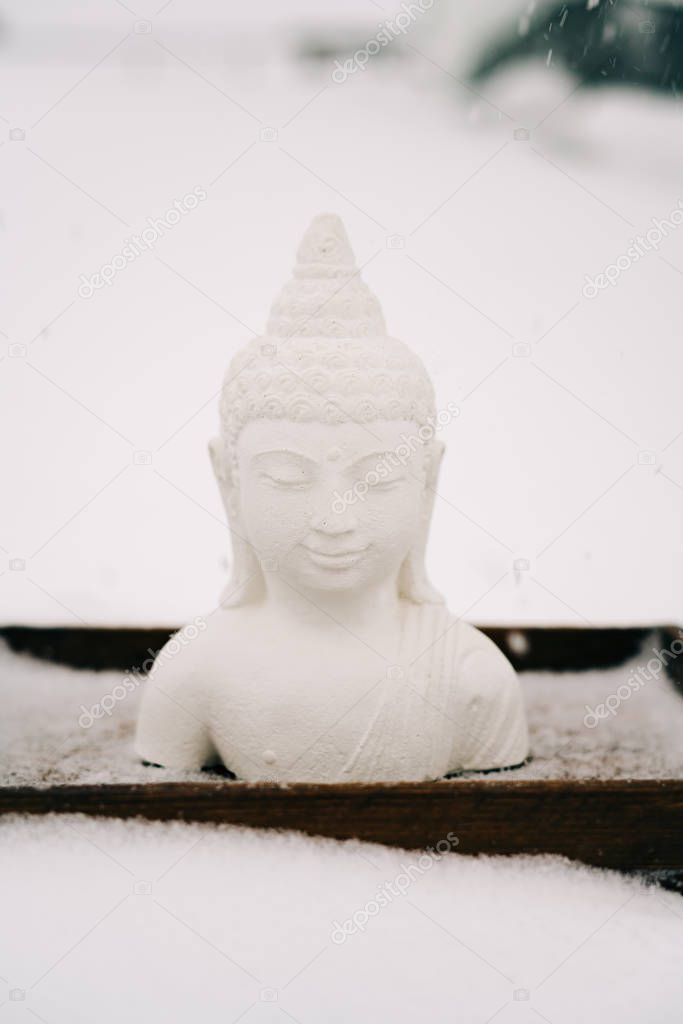 white Buddha figurine made of plaster on a wooden stand during a snowfall