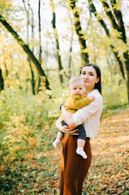 Genç bir anne, küçük mutlu oğlunu kollarında tutar sonbahar ağaçlarının arasında batan güneşin ışınlarında.