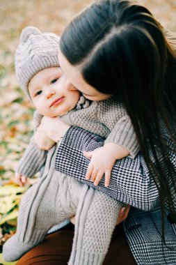 Bir anne, sonbahar mevsiminde, sonbahar yapraklarıyla kaplı bir düzlükte, parkta, sıcak bir tulum içinde, tatlı bir çocuğu kucağına alır.
