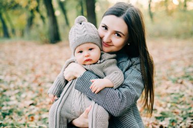 Bir anne, sonbahar mevsiminde, sonbahar yapraklarıyla kaplı bir düzlükte, parkta, sıcak bir tulum içinde, tatlı bir çocuğu kucağına alır.