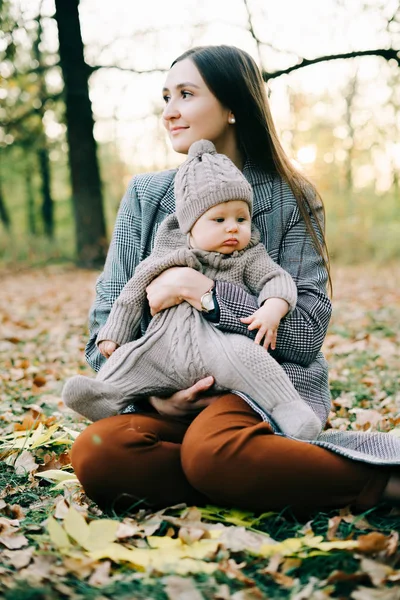 a mother holds a cute little boy in a warm jumpsuit in the Park in a clearing strewn with autumn leaves, in the autumn time