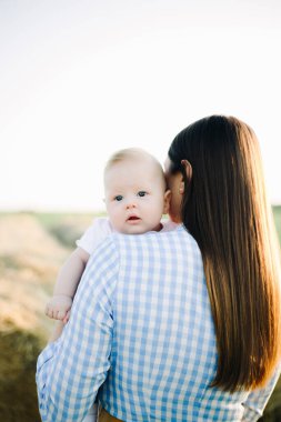 Mavi elbiseli genç bir anne sırtında küçük bir çocuğa dikilir ve yaz güneşinin altında omzunun üstünden kameraya bakar.