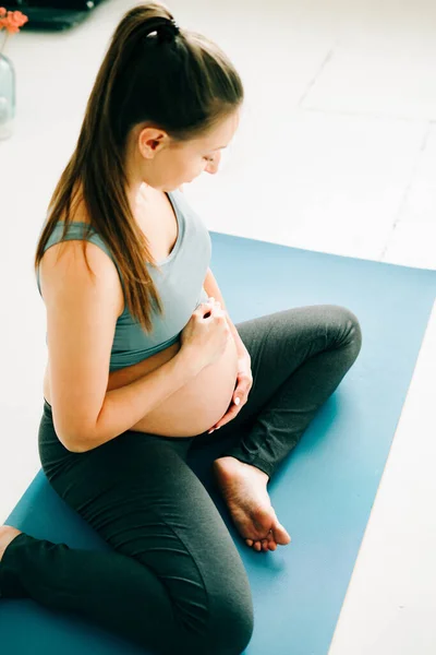 beautiful pregnant girl in sportswear sitting on a yoga Mat and stroking her belly