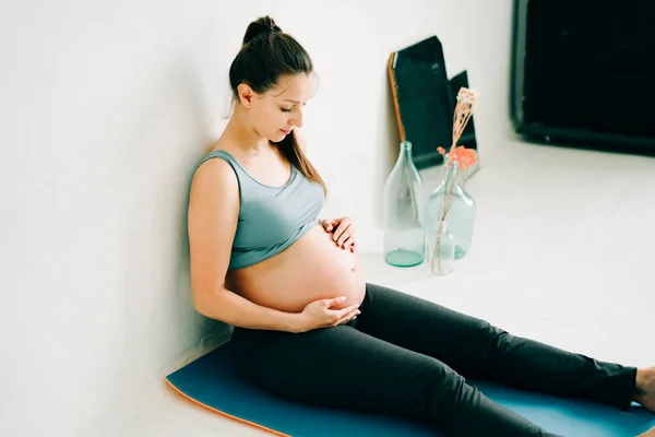 Spor kıyafetleri içinde genç ve güzel bir kız bir halıya oturup göbeğine sarılıyor.