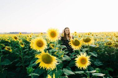 Siyah çiçekli bir elbise giyen genç ve güzel bir kız bir tarlada ayçiçeği çiçeklerinin arasında duruyor.