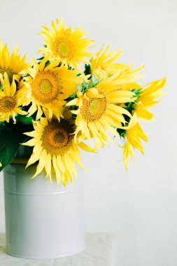 Masanın üzerinde beyaz bir masa örtüsüyle kaplı vazoda duran parlak sarı ayçiçeği çiçekleri.