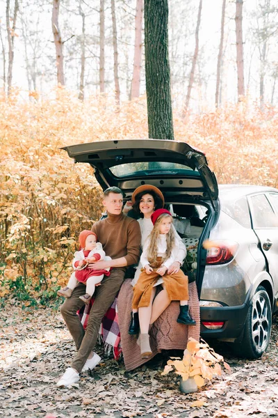 a happy family of four people in an autumn pine forest sit in the cozy trunk of their car covered with blankets. golden leaves