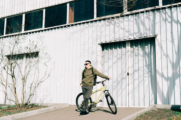 a young man in khaki sportswear holds a Bicycle standing next to him, against the background of a light hangar