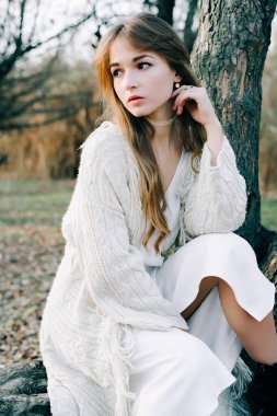 Açık beyaz elbiseli, siyah ayakkabılı genç, çekici bir kızın portresi. Resimli bir ağacın kökünde oturuyor.