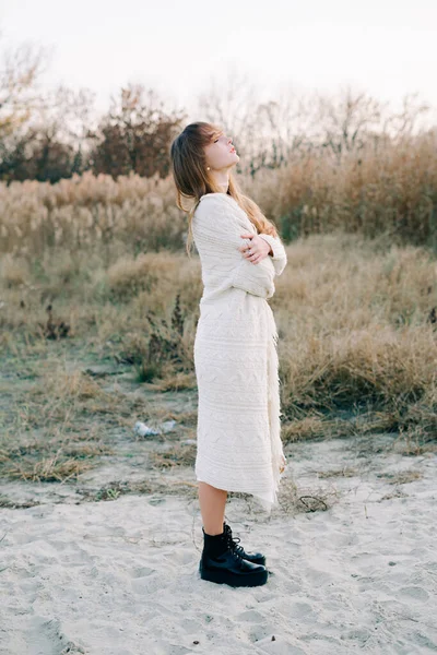 portrait of a young girl in a white dress, a warm knitted cardigan and black shoes among dry reeds and green bushes on the Bank of a river with white sand