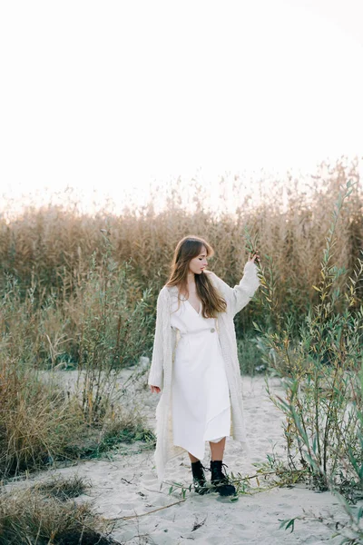 portrait of a young girl in a white dress, a warm knitted cardigan and black shoes among dry reeds and green bushes on the Bank of a river with white sand