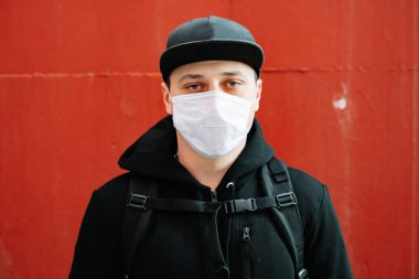 Siyah spor giyimli genç bir adam ve kırmızı duvarın yanında yüzünde tıbbi maske olan bir beysbol şapkası. Virüsten koruma