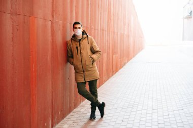 Kahverengi ceketli, yüzünde tıbbi maske olan genç bir adam sokakta, kırmızı duvarın yanında. Virüsten koruma