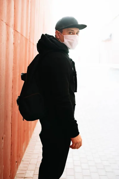 a young man in black sportswear and a baseball cap with a medical mask on his face near the red wall. protection from the virus