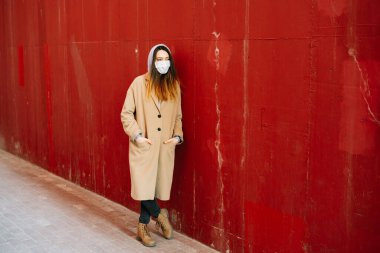 Caddede, kırmızı duvarın yanında, yüzünde tıbbi maske olan kahverengi paltolu genç bir kız. Virüsten koruma