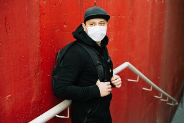 Siyah spor giyimli genç bir adam ve kırmızı duvarın yanında yüzünde tıbbi maske olan bir beysbol şapkası. Virüsten koruma