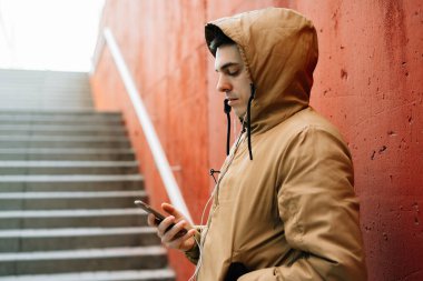 Caddede kırmızı duvarın yanında duran kahverengi ceketli genç bir adam akıllı telefonunu kullanıyor. sokak biçimi