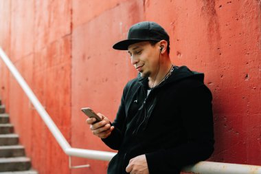Siyah spor giyim ve sokakta kırmızı duvarın yanında duran beysbol şapkalı genç bir adam akıllı telefonunu kullanıyor. sokak biçimi