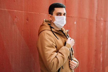 Kahverengi ceketli, yüzünde tıbbi maske olan genç bir adam sokakta, kırmızı duvarın yanında..