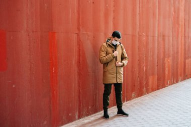 Kahverengi ceketli, yüzünde tıbbi maske olan genç bir adam sokakta, kırmızı duvarın yanında..