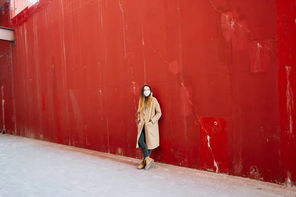 갈색 코트를 입고 얼굴에 의약품 마스크를 쓴 소녀, 빨간 벽 근처에서. — 스톡 사진