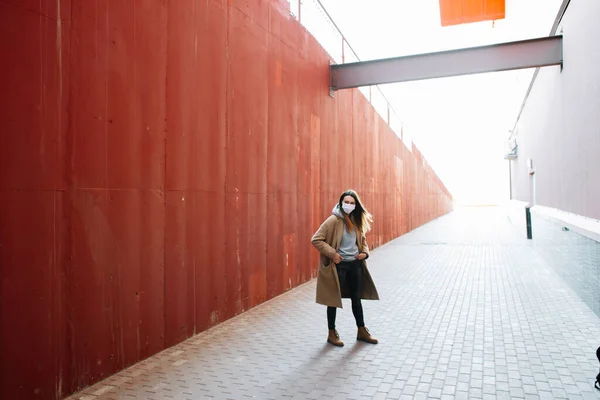 Una giovane ragazza in un cappotto marrone con una maschera medica sul viso per strada, vicino al muro rosso . — Foto Stock