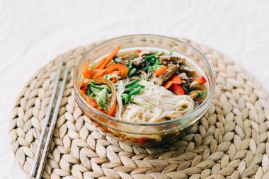 Bardakta Japon eriştesi çorbası, içinde vejetaryen malzemeler, küçük tabakta taze sebzeler, pirinç eriştesi, mantar ve miso ezmesi çorbası.