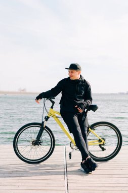 Siyah spor kıyafetli genç bir adam gölün yanındaki dubada duruyor ve yanında sarı bir bisiklet tutuyor.