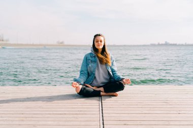 Genç ve güzel bir kız gölün yanındaki iskelede oturuyor ve meditasyon yapıyor.