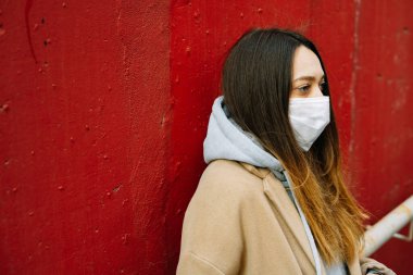 Caddede, kırmızı duvarın yanında, yüzünde tıbbi maske olan kahverengi paltolu genç bir kız. Virüsten koruma