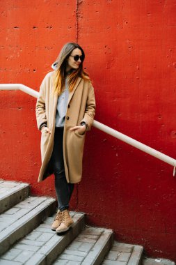 Caddede kırmızı duvarın yanında duran kahverengi ceketli ve gözlüklü genç bir kız. sokak biçimi