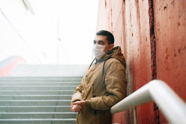 Kahverengi ceketli, yüzünde tıbbi maske olan genç bir adam sokakta, kırmızı duvarın yanında. Virüsten koruma