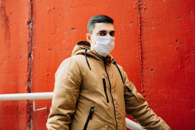 Kahverengi ceketli, yüzünde tıbbi maske olan genç bir adam sokakta, kırmızı duvarın yanında. Virüsten koruma
