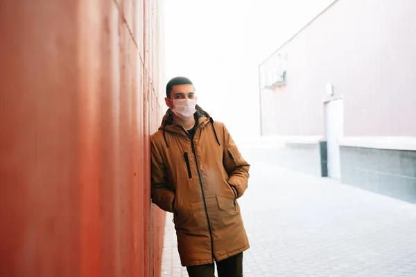 a young man in a brown jacket with a medical mask on his face on the street, near the red wall. protection from the virus