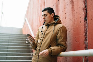 Caddede kırmızı duvarın yanında duran kahverengi ceketli genç bir adam akıllı telefonunu kullanıyor. .