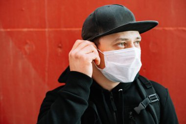 Siyah spor giyimli genç bir adam ve kırmızı duvarın yanında yüzünde tıbbi maske olan bir beysbol şapkası. Virüsten koruma