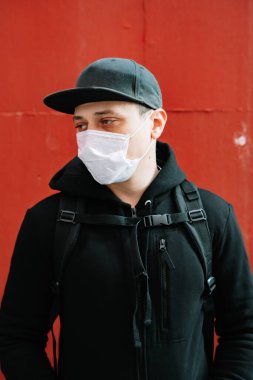 Siyah spor giyimli genç bir adam ve kırmızı duvarın yanında yüzünde tıbbi maske olan bir beysbol şapkası. Virüsten koruma