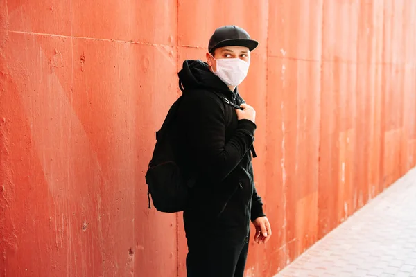a young man in black sportswear and a baseball cap with a medical mask on his face near the red wall. protection from the virus