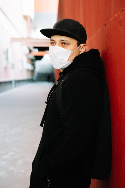A young man in black sportswear and a baseball cap with a medical mask on his face near the red wall. protection from the virus
