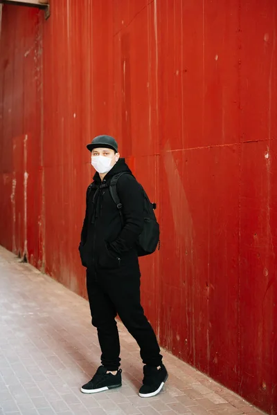 a young man in black sportswear and a baseball cap with a medical mask on his face near the red wall. protection from the virus