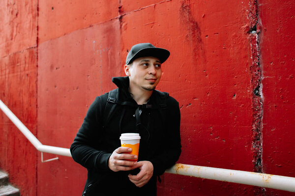 a young guy in black sportswear and a baseball cap on the street, standing near a red wall and drinking coffee .