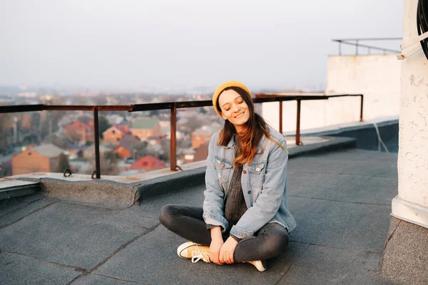 pretty white European woman is sitting on the roof of a multi-storey building in a blue denim jacket in gray pants,a yellow stylish hat.in the evening at sunset. lifestyle concept. trendy seasons