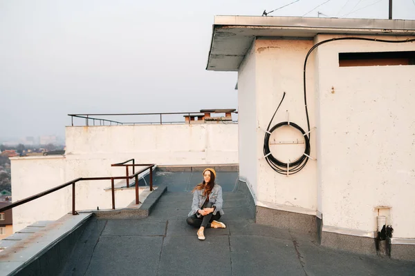 portrait white European woman is sitting on the roof of a multi-storey building in a blue denim jacket in gray pants,a yellow stylish hat.in the evening at sunset. lifestyle concept. trendy seasons