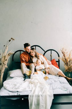 Moda kıyafetleri içinde mutlu ve şık bir aile, yatakta oturuyorlar, anne ve baba küçük oğullarına sarılıyorlar ve geniş bir stüdyoda birbirlerine gülümsüyorlar.