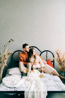 Moda kıyafetleri içinde mutlu ve şık bir aile, yatakta oturuyorlar, anne ve baba küçük oğullarına sarılıyorlar ve geniş bir stüdyoda birbirlerine gülümsüyorlar.