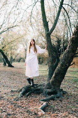 Açık beyaz elbiseli ve siyah ayakkabılı çekici sarışın bir kızın portresi bir ağacın kökünde duruyor.