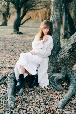 Açık beyaz elbiseli ve siyah ayakkabılı çekici sarışın bir kızın portresi bir ağacın kökünde duruyor.