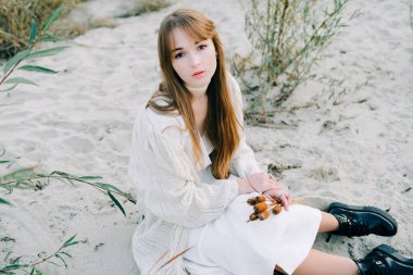 Beyaz elbiseli genç bir kızın portresi, kalın bir hırka ve kuru sazlıkların arasında siyah ayakkabılar ve beyaz kumlu bir nehrin kıyısında yeşil çalılar.